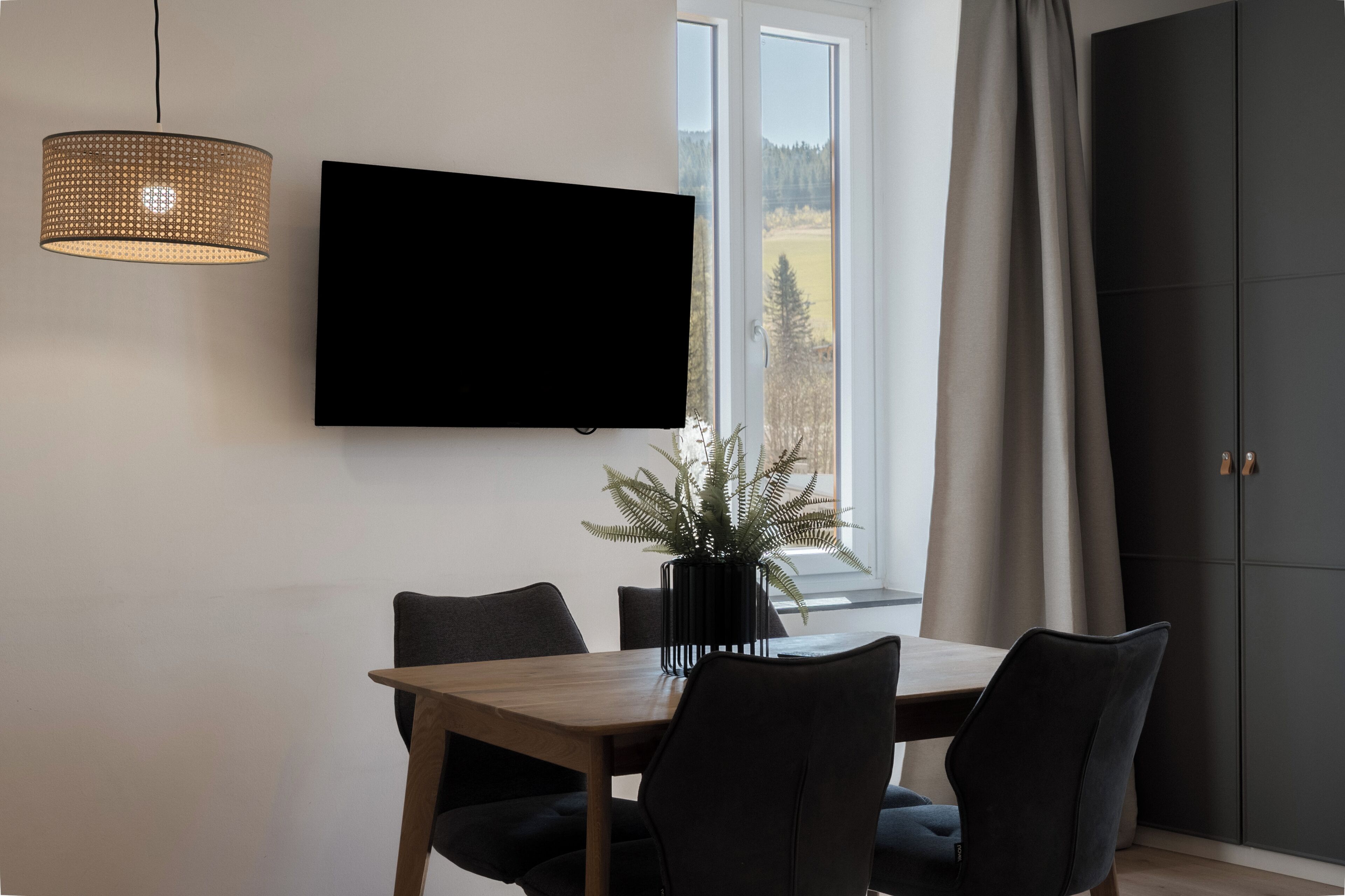 A modern dining area with a flat-screen TV, a woven pendant light, and a potted plant.