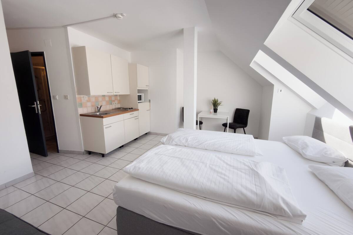 A modern bedroom with a large bed, a small desk, and a kitchenette in the background.