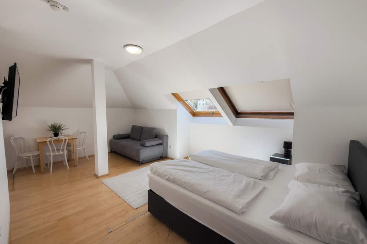 A modern loft bedroom with a skylight, a sofa, a dining table with chairs, and a small desk.