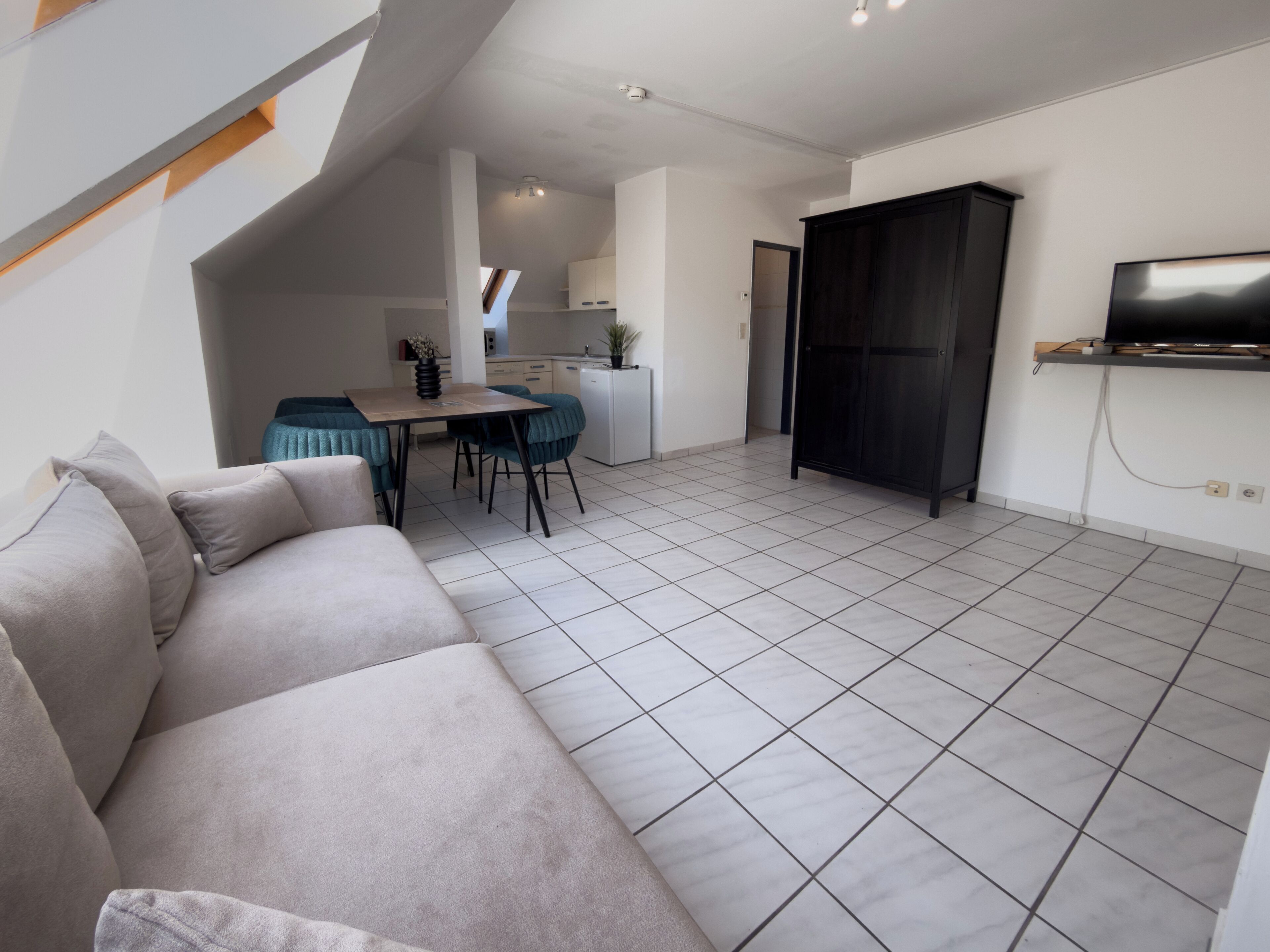 Een moderne woonkamer met een bank, een eethoek met stoelen, een keuken op de achtergrond en een tv aan de muur.