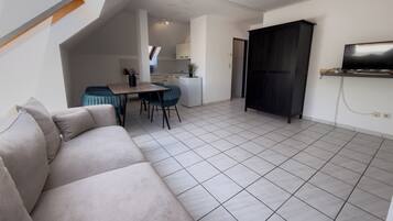 Een moderne woonkamer met een bank, een eethoek met stoelen, een keuken op de achtergrond en een tv aan de muur.