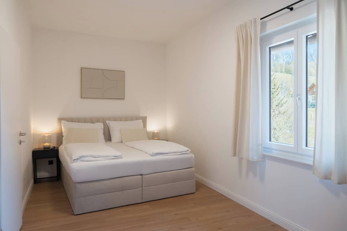A bedroom with a bed, bedside tables, a window with curtains, and a wall-mounted artwork.