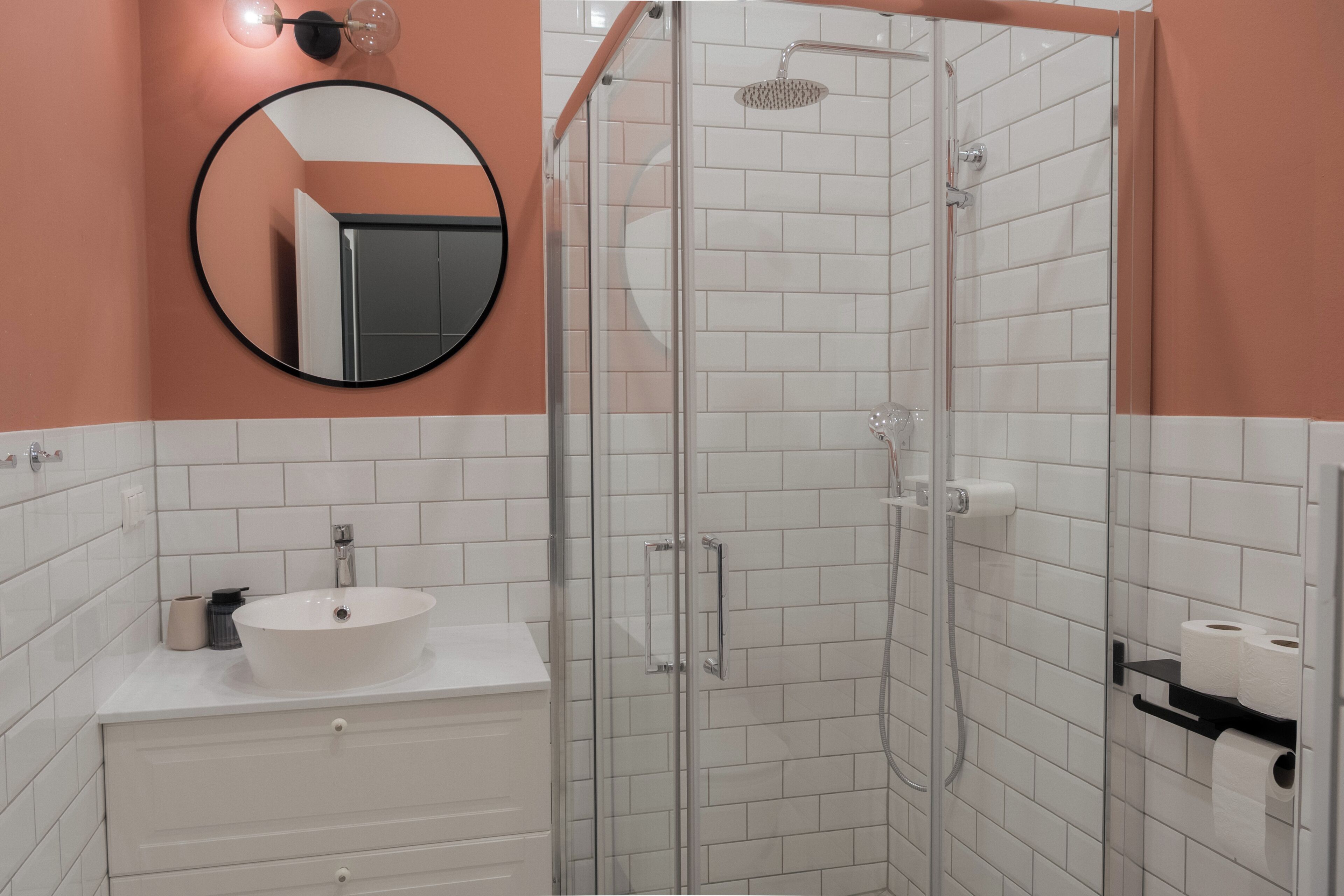 Un baño con un lavamanos blanco, cabina de ducha de vidrio, espejo redondo y dispensador de papel higiénico.