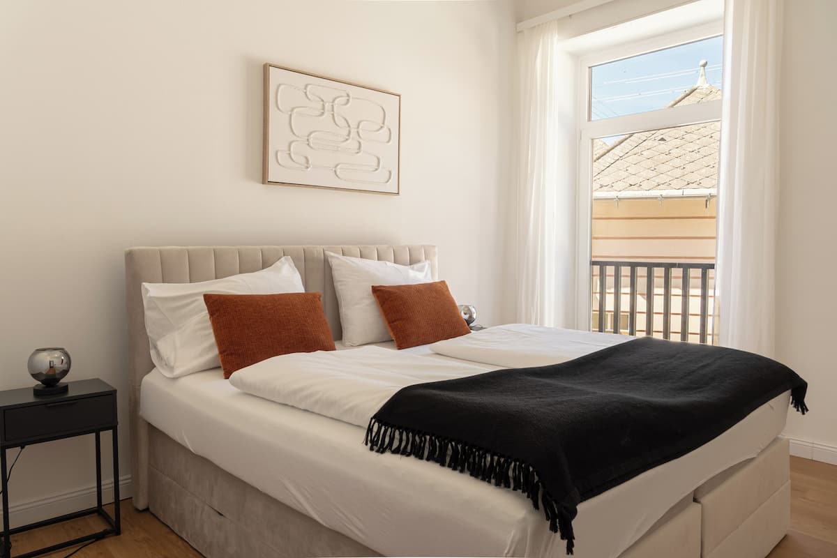 A bedroom with a bed, pillows, a bedside table, a wall art piece, and a window with a view of a building.