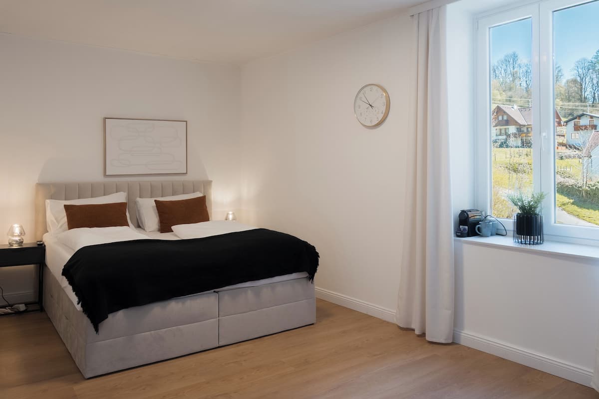 A bedroom with a bed, a clock, a window with a view of houses, and a bedside table with a lamp.