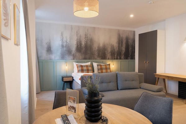 A modern hotel room with a sofa, a dining table, a vase, and a wall with a forest mural.