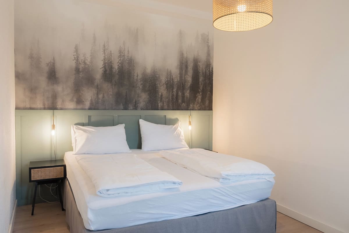 A neatly made bed with white linens, a headboard with wall-mounted lights, and a forest-themed wall mural.
