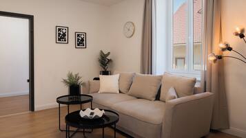 A modern living room with a beige sofa, a small black side table, a potted plant, and a wall clock.