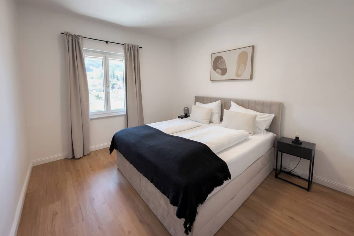 A bedroom with a bed, a nightstand, a window with curtains, and a wall-mounted artwork.