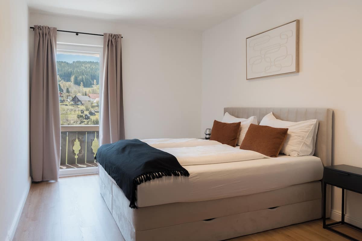 A bedroom with a bed, a window with curtains, a bedside table, and a wall-mounted picture.