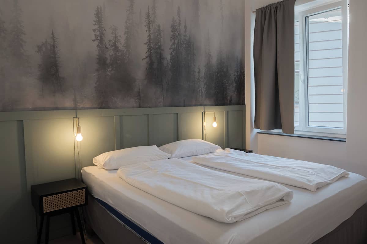 A bed with white linens, a headboard with wall-mounted lights, and a window with curtains.
