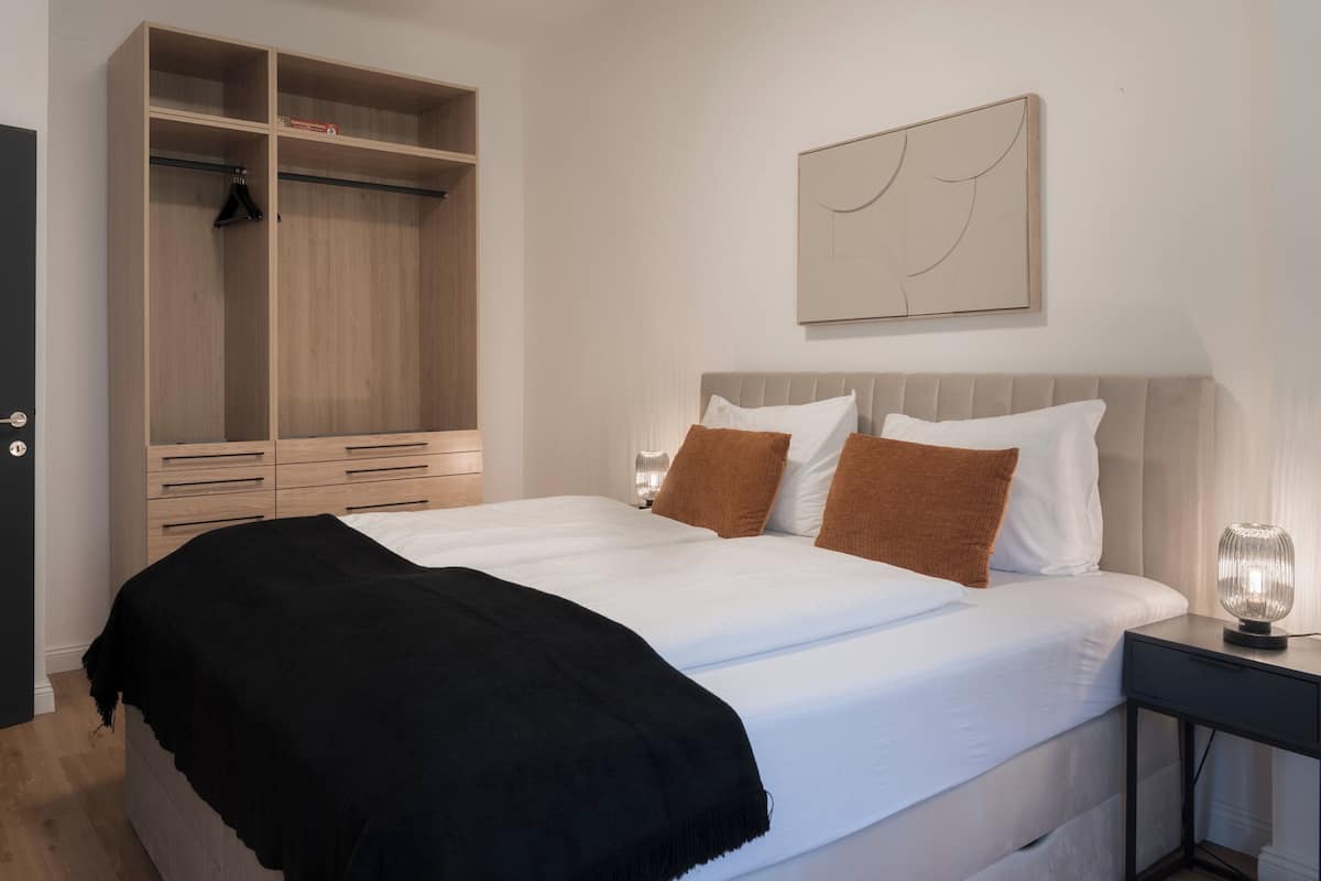 A bedroom with a bed, a wardrobe, a bedside table with a lamp, and a wall-mounted artwork.