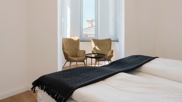 A bedroom with a bed, two armchairs, a small round table, and a window with a view of buildings. - Aparthotel Hirschenhof (Spital am Semmering)