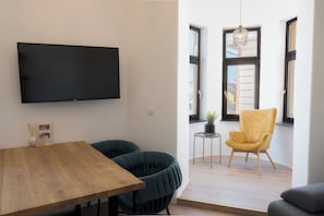 A modern living room with a flat-screen TV, a wooden dining table, two dark armchairs, a yellow armchair, and a small side table with a plant.