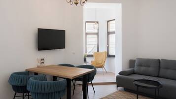 A modern living room with a sofa, a dining table with chairs, a TV mounted on the wall, and a chandelier.