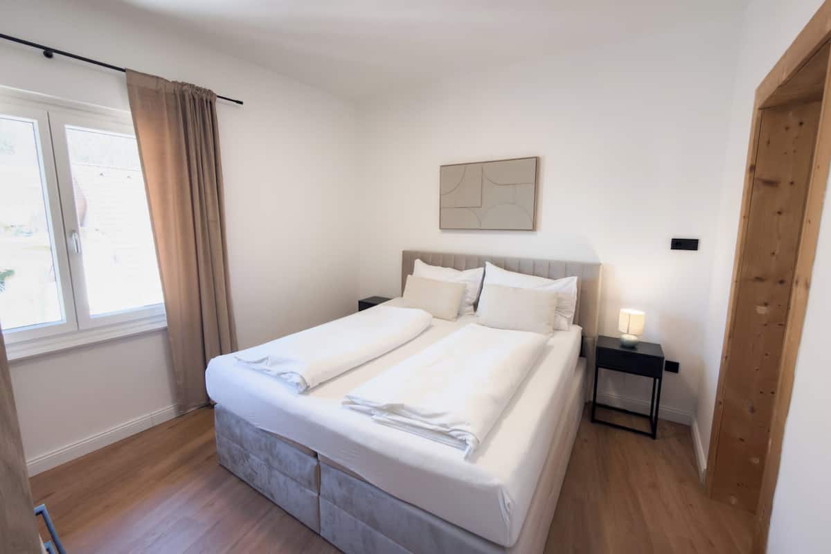 A bedroom with a bed, a nightstand, a lamp, a window with curtains, and a wooden door.