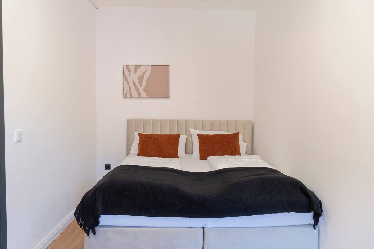 A bedroom with a bed, two pillows, a blanket, a headboard, and a wall-mounted artwork.