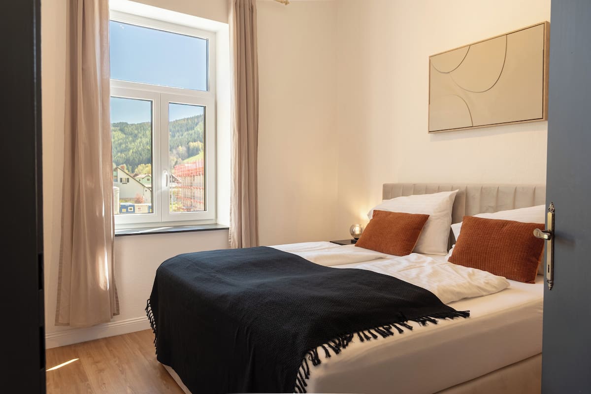 A bedroom with a bed, a window with curtains, a bedside table, and a wall-mounted artwork.