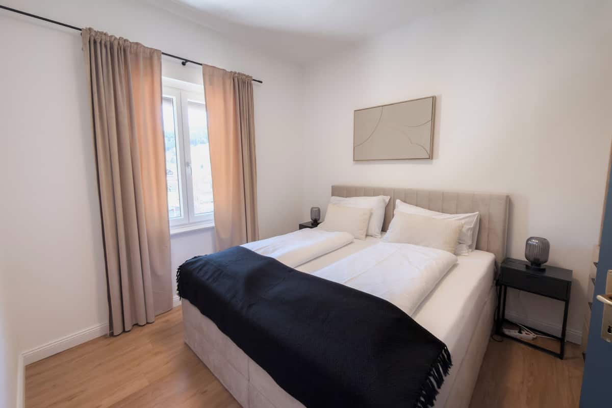 A double bed with a black throw blanket, a bedside table with a lamp, and a window with curtains.