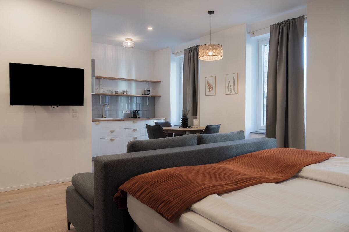 A modern hotel room with a bed, a sofa, a desk, and a kitchenette.
