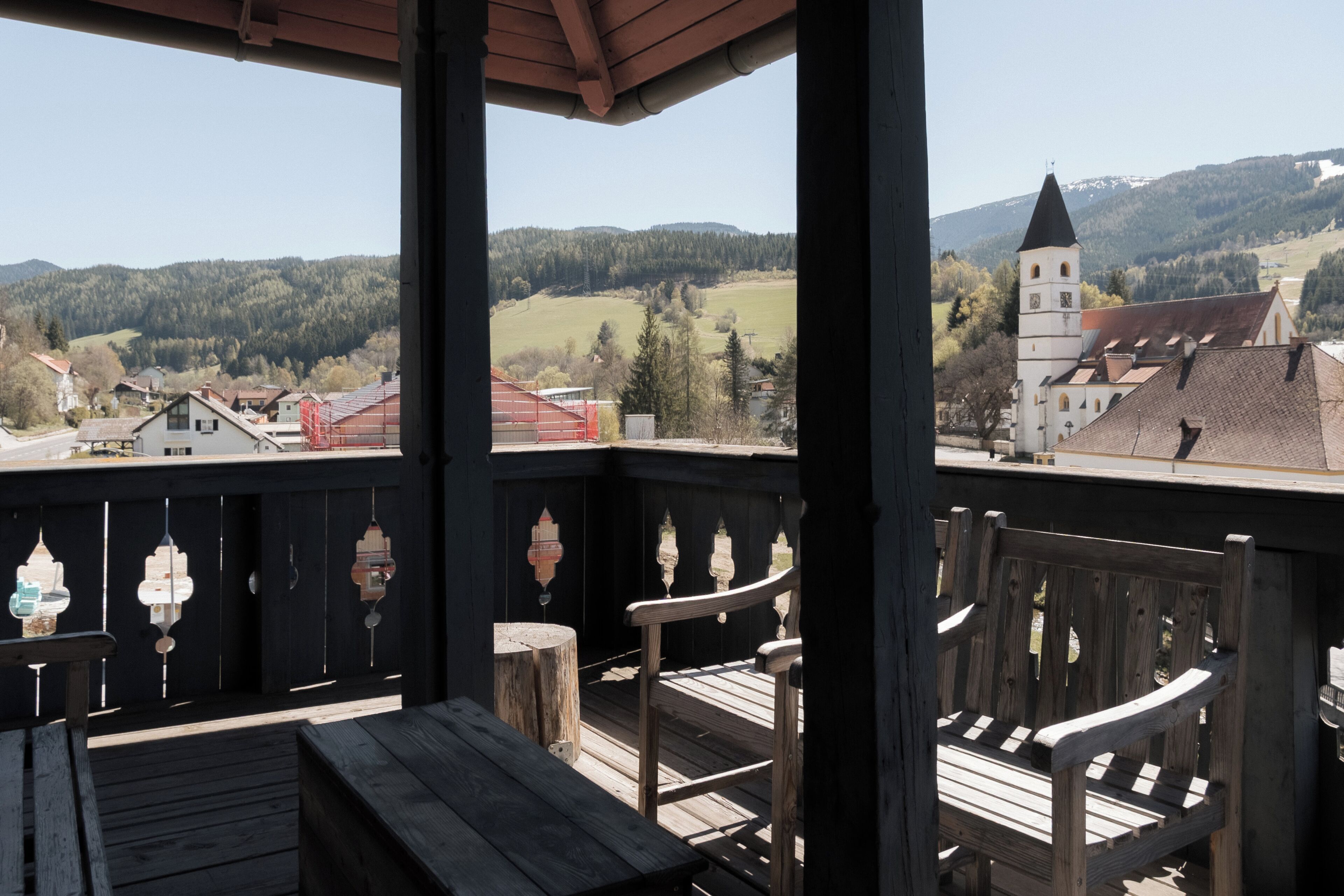 A wooden balcony with a bench, a view of a town with a church, and a mountain in the background.