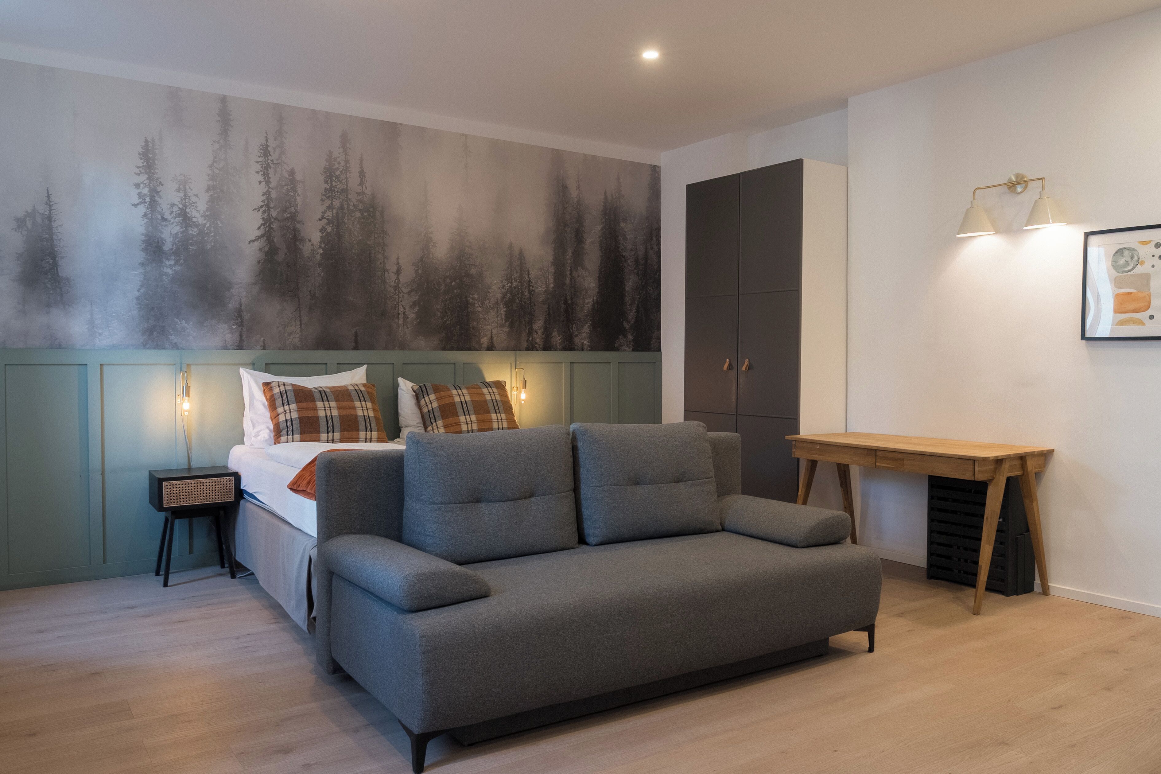 A modern bedroom with a bed, grey sofa, wooden desk, and a forest mural on the wall.