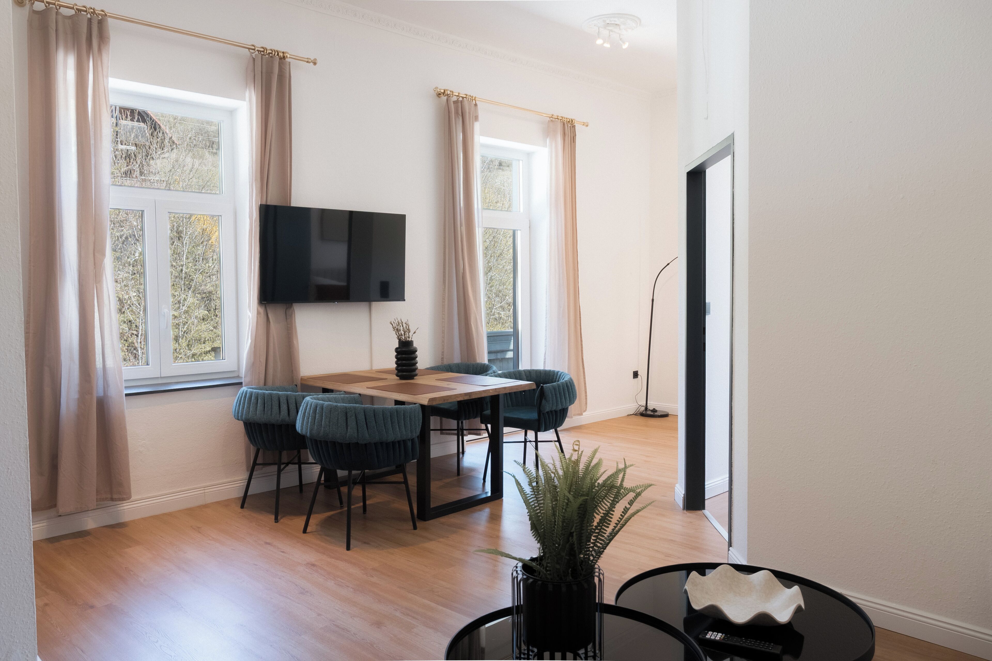 A modern living room with a wooden dining table, two chairs, a flat-screen TV, and a large window with curtains.