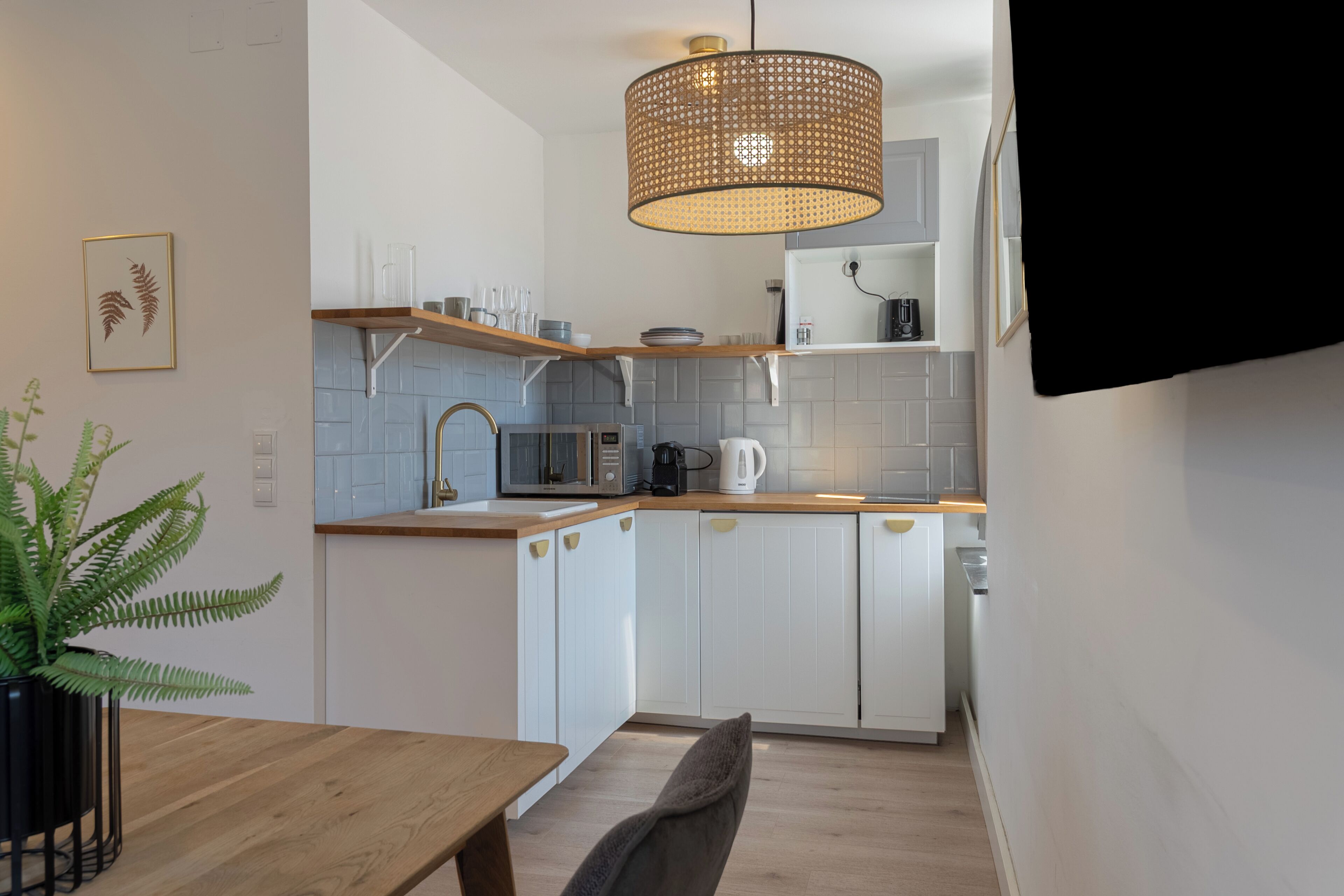 Une cuisine moderne avec des meubles blancs, un plan de travail en bois, un luminaire suspendu en rotin et une fougère en pot.