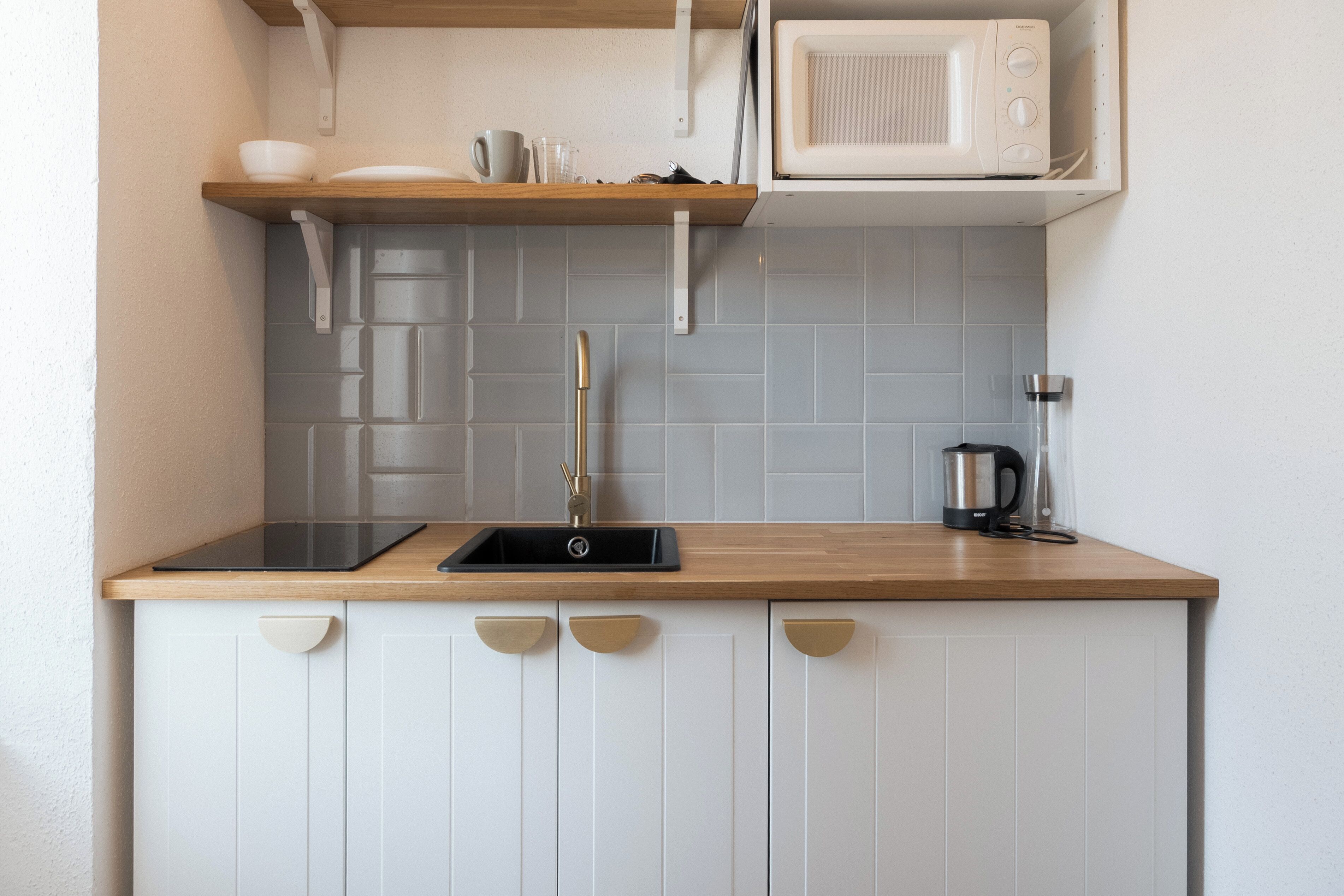A kitchen with a wooden countertop, white cabinets, a sink, a microwave, and a kettle.