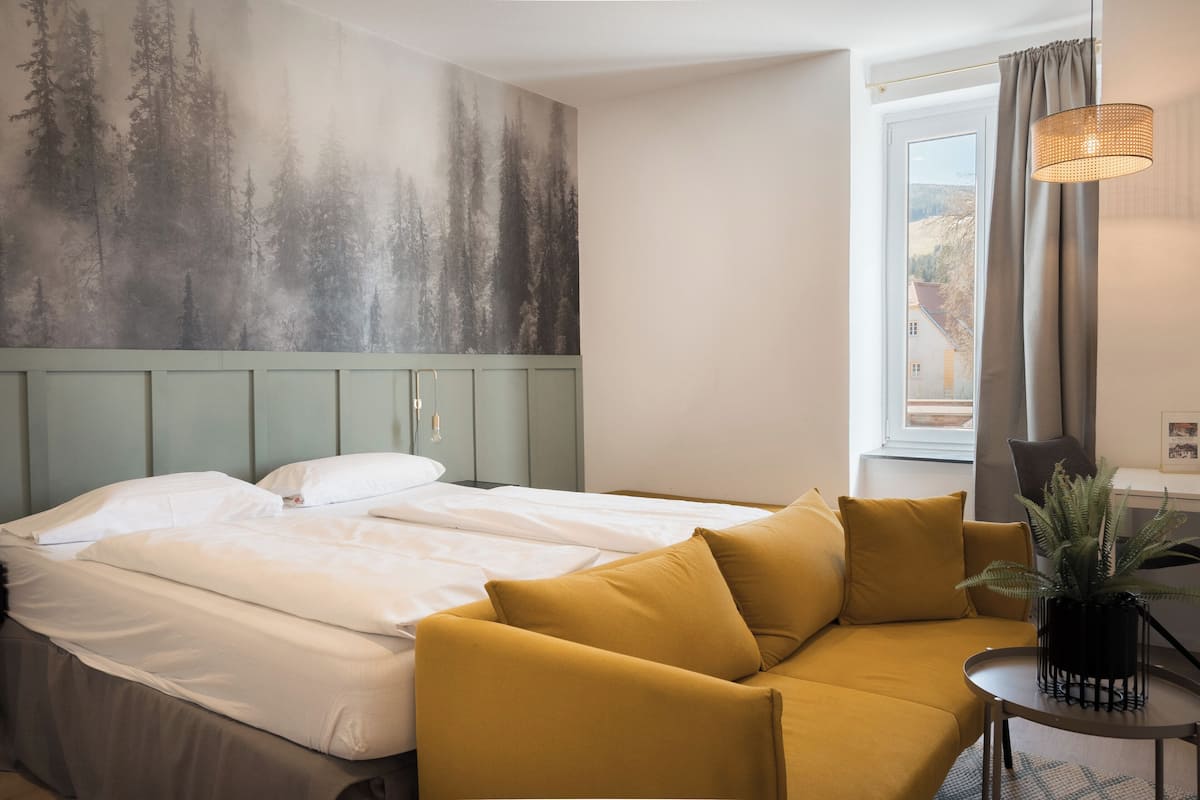 A bedroom with a bed, a sofa, a window with curtains, a wall with a forest mural, and a hanging light fixture.