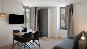 A modern living room with a sofa, dining table, chairs, a flat-screen TV, and a large window with curtains. - Aparthotel Hirschenhof (Spital am Semmering)