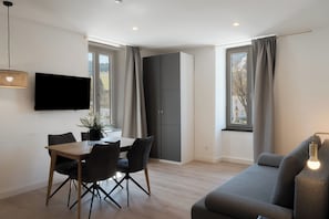 Un salon moderne comprenant un canapĂ©, une table Ă manger, des chaises, un tĂ©lĂ©viseur Ă Ă©cran plat et une grande fenĂȘtre avec des rideaux.