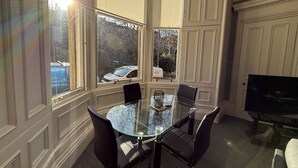 Ein Zimmer mit einem Glastisch, vier Stühlen, einem Fernseher und großen Fenstern mit Blick auf geparkte Autos und Bäume.