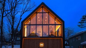 Una cabaña moderna de madera con ventanas amplias, un interior visible con una cama y un cartel que indica un restaurante.