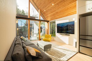 A modern living room with a sofa, ottoman, and a flat-screen TV mounted on the wall. - Echowoods.  Luxe. Fireplace. Onsen (Hakuba Village, Hokujo)