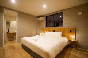 Une chambre dâhĂŽtel avec un grand lit, une tĂȘte de lit en bois, une fenĂȘtre, une table de chevet avec une lampe et une salle de bain visible Ă travers une porte ouverte.