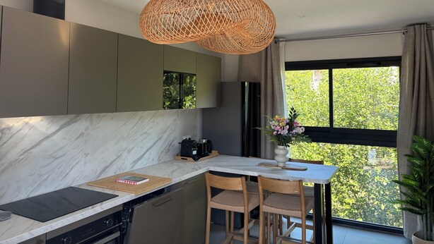 Een moderne keuken met een ingebouwde kookplaat, een eethoek met een tafel en stoelen, en een groot raam met uitzicht op groen.