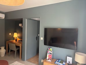 A room with a flat-screen TV mounted on the wall, a wooden table with a lamp, a chair, and a small table with books and a brochure. - T2 Puy de dôme Chic, Cosy & Idéalement Situé (Clermont-Ferrand)