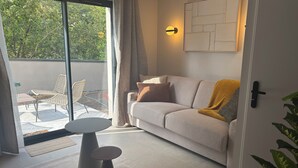 Een moderne woonkamer met een bank, een klein rond tafeltje, een glazen deur die leidt naar een buiten zitgedeelte en een wandlamp.