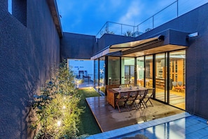 Eine moderne Außenpatio mit Holztisch und -stühlen, einem kleinen Garten und einem wintergartenartigen Essbereich.