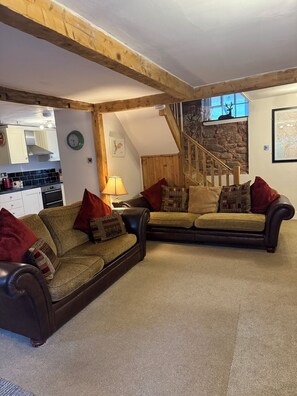 Ein gemütliches Wohnzimmer mit zwei dunkelbraunen Ledersofas, einer Holztreppe, einer Küche im Hintergrund und einer Steinwand.