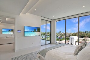 Een moderne slaapkamer met een groot bed, een televisie en een balkon met uitzicht op een strand en parkeerplaats.