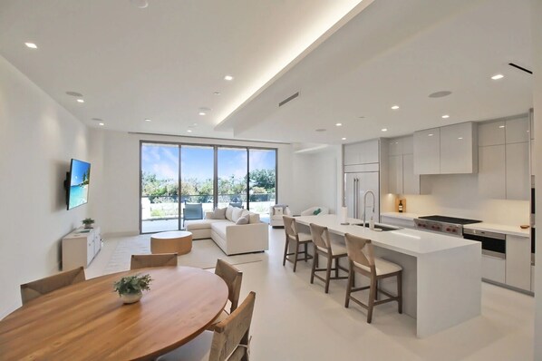 Een moderne keuken met een centraal eiland, een eethoek met een houten tafel en een zithoek met een bank en televisie.