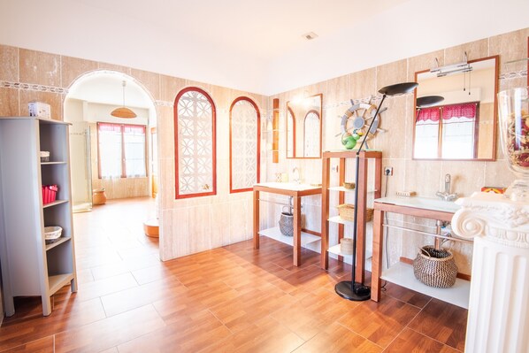 A well-lit room with a wooden floor, a white vanity with a mirror, a shelf with baskets, and a doorway leading to another room.