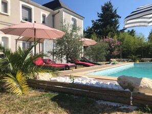 A pool area with lounge chairs and umbrellas, surrounded by a house and greenery.