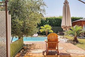 Um deck de madeira com cadeira e mesa, piscina e cerca verde.