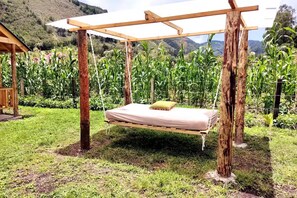 Un hamac tendu sous une pergola en bois, dans un jardin oĂč poussent des plantes de maĂŻs, avec une chaĂźne de montagnes en arriĂšre-plan.