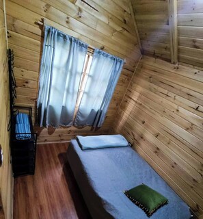 Une petite pièce de cabane en bois, avec un lit simple, une fenêtre avec des rideaux et une petite table accompagnée d’une chaise.