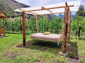 Un lit-balançoire en bois, doté d’un matelas et d’un oreiller, entouré d’une zone herbeuse et d’un champ de maïs, avec une montagne en arrière-plan.