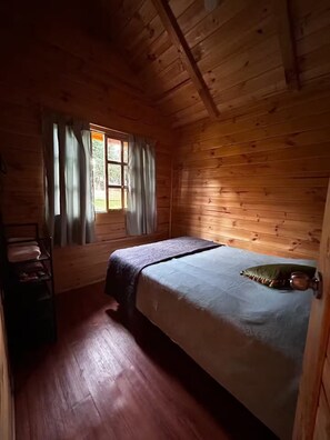 Una habitación de cabaña de madera con una cama, un ventanal con cortinas y un estante con toallas.